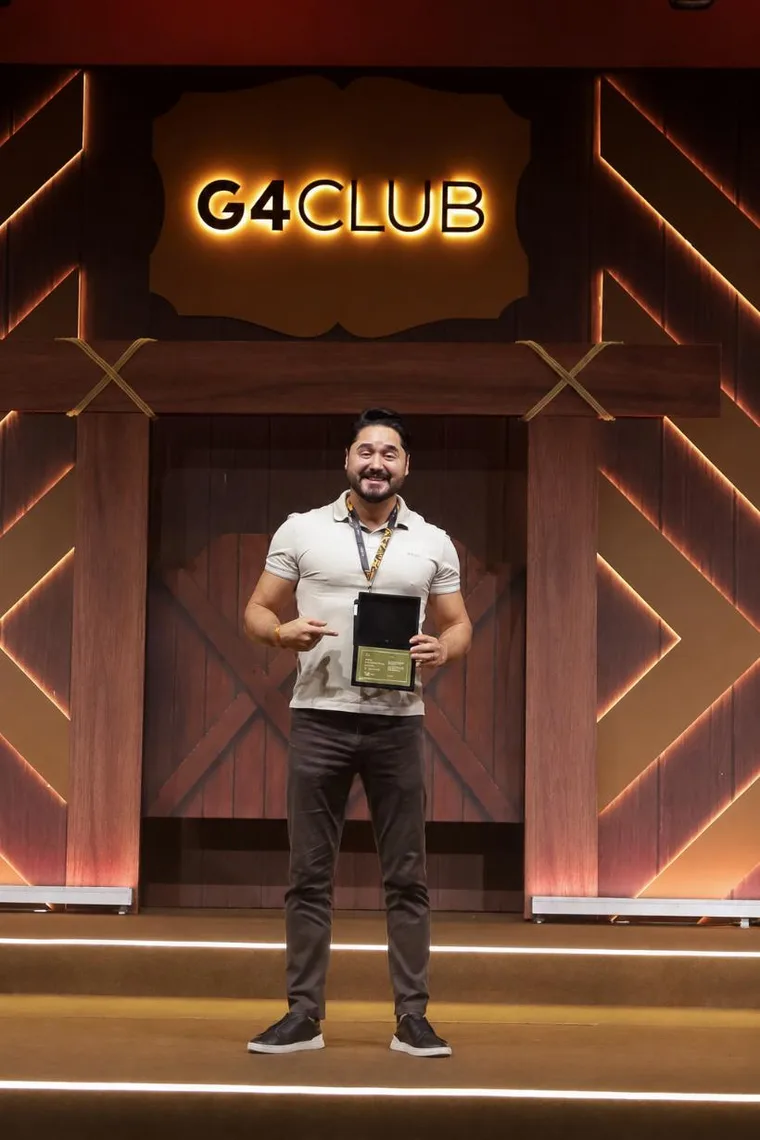 Alex Pinhol no palco do G4 Club segurando a placa do reconhecimento Eagle,
              sob o letreiro iluminado do clube