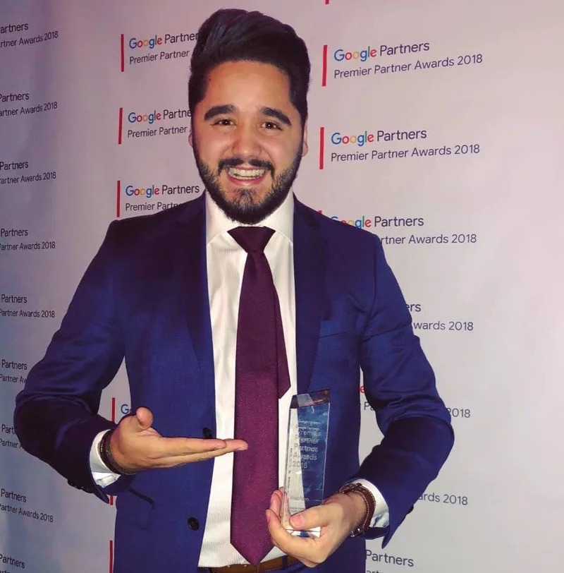 Alex Pinhol segurando o troféu do Google Partners Premier Partner Awards 2018, diante do painel do evento